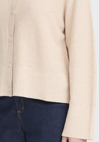 Cardigan bej cu model cu dungi și nasturi în față, mâneci late, asortat cu blugi albastru închis cu cusături galbene.