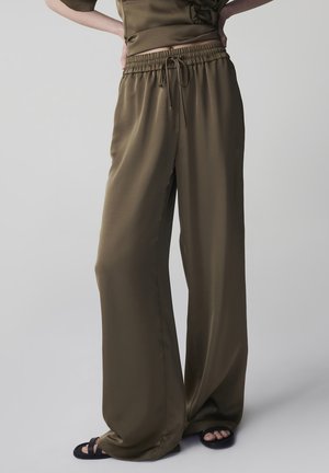 Pantalones de satén verde oliva de corte holgado con cintura elástica y cordón, usados con sandalias negras tipo slide, en una persona de pie.