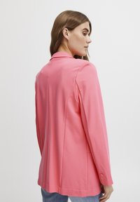 Rožs blazeris, izgatavots no gludas auduma, ar pielāgotu piegriezumu, izgriezumu ar uzliekamu apkakli, garām piedurknēm un pogu akcentiem uz piedurknēm.