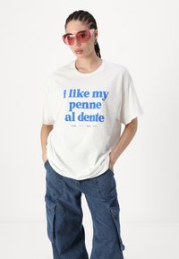 Balta medvilninė marškinėliai su mėlynu užrašu "Man patinka mano penne al dente." Derinami su mėlynomis džinsinėmis krovinių kelnėmis. Modelis dėvi raudonus akinius nuo saulės.