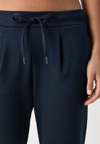 Marineblauwe broek gemaakt van zachte stof, met voorplooien, een taille met trekkoord en zilveren metalen uiteinden, en zijzakken.