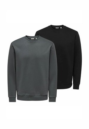 Junger Mann steht mit den Händen hinter dem Rücken, trägt einen schwarzen Pullover, hellgraue Jeans und schwarze Sneakers mit weißen Sohlen.