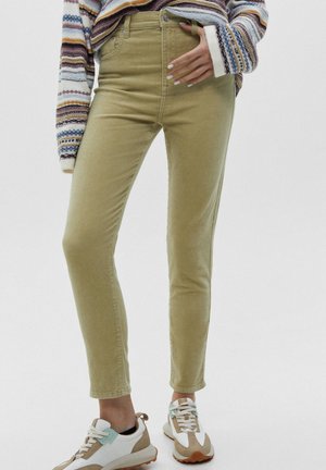 Personne portant un pantalon beige slim en velours côtelé, un pull rayé multicolore et des baskets blanches et beiges, debout sur un fond blanc.