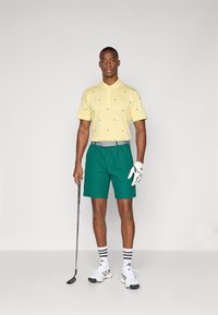 Polo tričko ve světle žluté barvě s zelenými vzory listů, spárované s tmavě zelenými šortkami. Bílé sportovní boty a doplněk v podobě golfové hole.