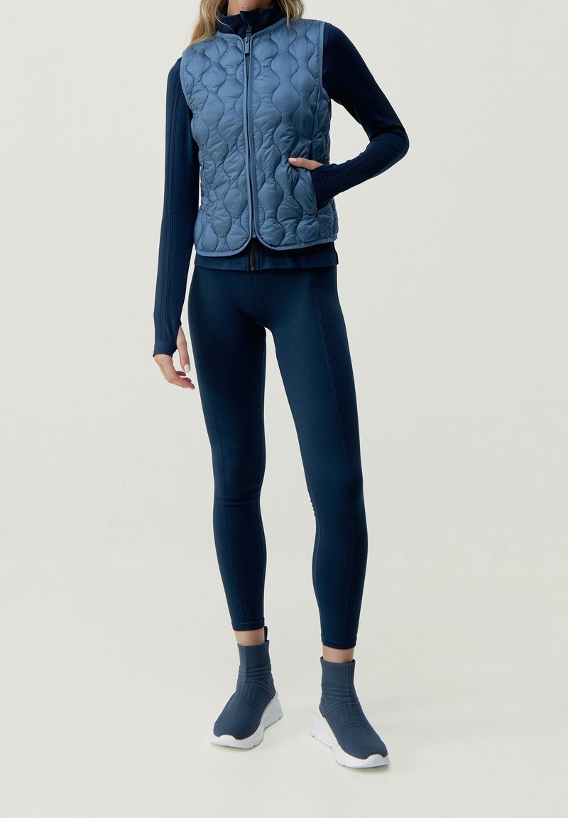 Chaleco acolchado azul con cuello alto, diseño ajustado y bolsillos laterales, sobre una camiseta de manga larga azul marino y leggings a juego. Zapatillas con suela blanca.