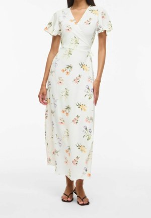 Femme portant une robe portefeuille blanche à fleurs avec des manches courtes flottantes et des sandales noires à brides, debout sur un fond blanc.