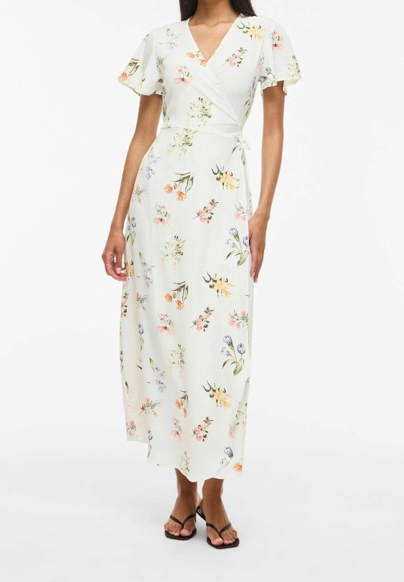 Femme portant une robe portefeuille blanche à fleurs avec des manches courtes flottantes et des sandales noires à brides, debout sur un fond blanc.