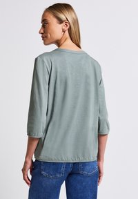Top verde chiaro a maniche 3/4 con collo rotondo e polsini elasticizzati, indossato con jeans blu scuro. Tessuto morbido e design dal fit rilassato.