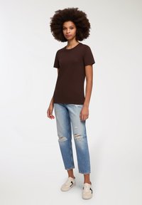 Brązowa koszulka z krótkim rękawem połączona z jasnoniebieskimi dżinsami w stylu distressed i beżowymi sneakerami. Stylizacja charakteryzuje się prostym wzornictwem i luźnym krojem.