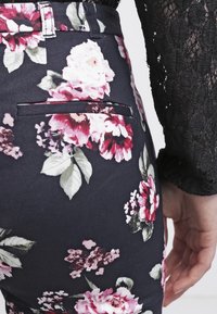 Květinové kalhoty s tmavým pozadím, které mají růžové a červené květy, slim fit střih a detail zadní kapsy. Měkký materiál.