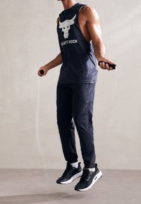 Mustat hihattomat tank topit, joissa on harmaa härkäkuvio, mustat urheiluhousut ja mustat lenkkarit, pitäen hyppynarusta ilmaan neutraalia taustaa vasten.