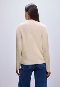 Jersey de color crema, texturizado, con mangas largas, cuello redondo y un corte holgado, combinado con jeans azul.