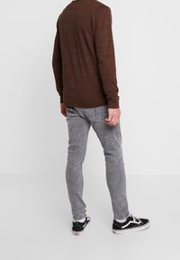 Hombre vistiendo una camisa de manga larga marrón oscuro, jeans gris claro, calcetines blancos y zapatillas negras de perfil bajo, de pie con la espalda hacia la cámara.