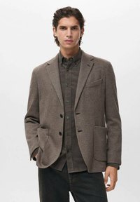Blazer marrone a spina di pesce con revers a ramponi, due tasche frontali e bottoni neri. Indossato sopra una camicia nera con bottoni e pantaloni neri.