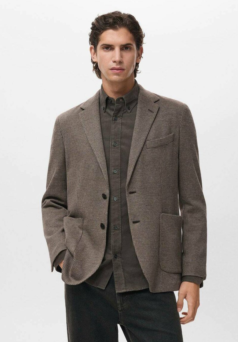 Blazer marrone a spina di pesce con revers a ramponi, due tasche frontali e bottoni neri. Indossato sopra una camicia nera con bottoni e pantaloni neri.