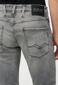 Κοντινή λήψη της πίσω τσέπης γκρίζων τζιν Replay με μαύρη ραφή σχεδίου και μαύρο δερμάτινο λογότυπο στη ζώνη μέσης.