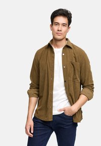 Camisa de pana marrón con botones en la parte delantera, un bolsillo en el pecho y mangas remangadas, combinada con una camiseta blanca y jeans oscuros.