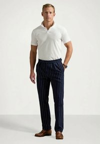 Άνδρας στέκεται με το ένα χέρι στην τσέπη, φορώντας λευκό πόλο μπλουζάκι, ναυτικές ραβδωτές παντελόνες, καφέ παπούτσια και ρολόι στο αριστερό του χέρι.