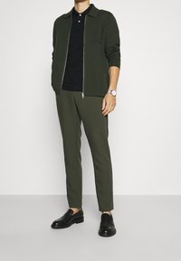 Olivgrön zip-up kofta över en svart polo, kombinerat med olivgröna tapered byxor och svarta loafers. Silverur på handleden.