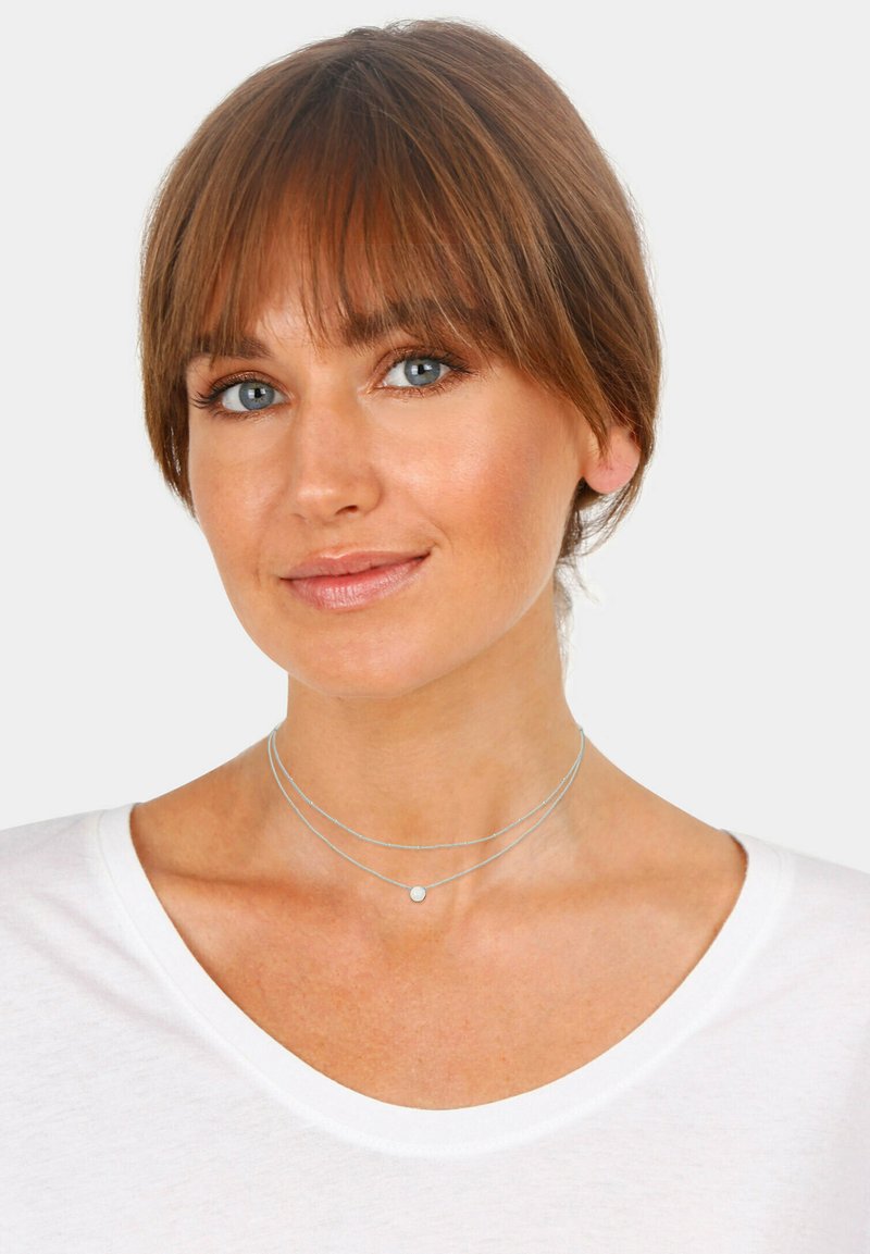Silver layered choker necklace featuring three strands and a small circular pendant, worn over a white shirt with a wide neckline.