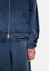 Άτομο που φοράει φθαρμένο φούτερ με φερμουάρ σε ναυτικό μπλε χρώμα και αντίστοιχο παντελόνι, με το χέρι χαλαρό στο πλάι, ορατά φθαρμένα άκρα στα μανίκια και στο τελείωμα.