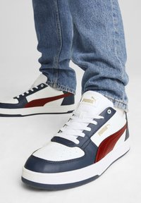 Las zapatillas Puma cuentan con una parte superior de cuero blanco con acentos en azul marino y rojo, una puntera perforada, cordones planos y una suela de goma texturizada.