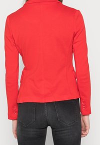 Femme portant un blazer rouge ajusté avec des manches boutonnées et un jean noir, vue de dos sur un fond uni.