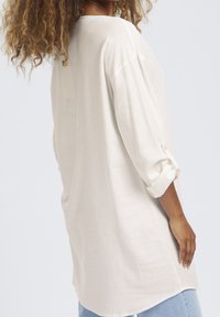 Jasnobeżowa lniana koszula o luźnym kroju, z rękawami do połowy przedramienia i mankietami zapinanymi na guziki. Gładka tekstura i subtelny układ materiału.