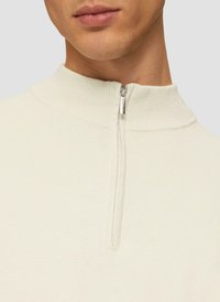 Trekker in lichtcrème, met een hoge hals, geribbelde textuur en een zilveren rits aan de voorkant, met een minimalistisch ontwerp.