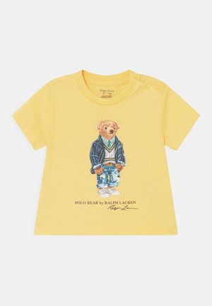 Gul kortærmet T-shirt med en bjørn i en stribet blazer, hvid sweater, blå jeans og sneakers. Illustration med brandtekst nedenfor.