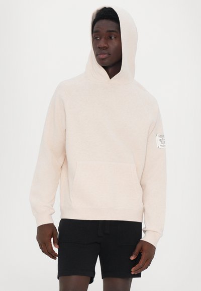 Hellbeige Kapuzenpullover mit Kängurutasche, verstellbarer Kapuze und einem Etikett am linken Ärmel. Der Stoff wirkt weich und strukturiert.