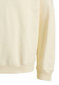 Felpa color crema con una texture liscia, polsini e orlo a coste, e dettagli di cucitura minimi. Presenta un piccolo etichetta sul lato.