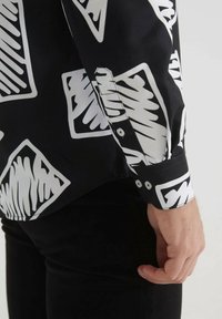 Chemise noire avec des motifs abstraits blancs, poignets à boutons et une texture de tissu lisse. Main visible reposant sur un pantalon noir.