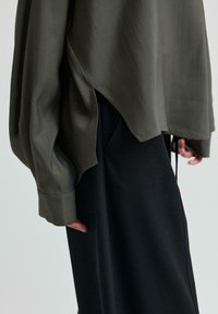 Blusa oversize verde oliva con maniche lunghe, texture liscia e aperture laterali, abbinata a un pantalone nero aderente.