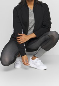 Musta urheilu takki, jossa on sivustripit, harmaa T-paita alla, teksturoitu musta ja hopea kuvioitu leggingsi, ja valkoiset lenkkarit.