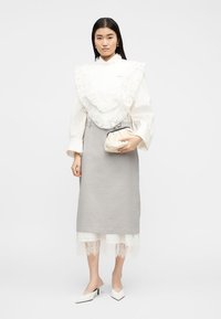 Femeie purtând o bluză albă cu volane din dantelă, fustă midi gri peste o fustă intermediara albă cu margini de dantelă, ținând o poșetă mică crem, pantofi cu toc albi.