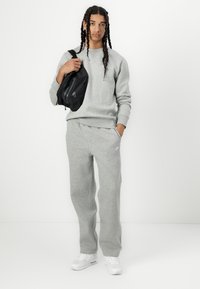 Grå genser og matchende joggebukser laget av mykt stoff. Modellen holder en svart, buet skulderveske med glidelåslomme. Hvite joggesko.