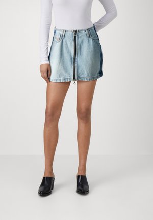 Femme portant une mini-jupe en denim bleu clair avec fermeture éclair à l'avant, un top blanc à manches longues et des chaussures à talons noires à enfiler, debout sur un fond uni.