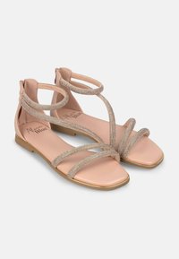 Sandalias planas con una base nude, que cuentan con varias correas brillantes y soporte en el tobillo. La suela es minimalista con un acento dorado alrededor del borde.