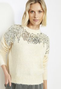 Kreemivärvi kampsun, valmistatud pehmest kootud materjalist, millel on õlgadel hõbedased helmestega mustrid, soonikservade ja varrukapitsidega.