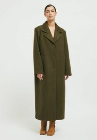 Olive green long coat with a notched collar, five buttons, two side pockets, and a straight silhouette, paired with brown ankle boots.