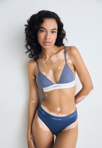 Ensemble de lingerie Calvin Klein comprenant un bralette gris côtelé avec une bande inférieure blanche et un slip bleu assorti, tous deux présentant un accent logo.