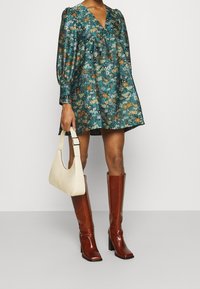 Tealfarbenes Kleid mit Blumenmuster, Puffärmeln, V-Ausschnitt und tailliertem Schnitt. Kombiniert mit braunen kniehohen Stiefeln mit Absatz und einer weißen Handtasche.