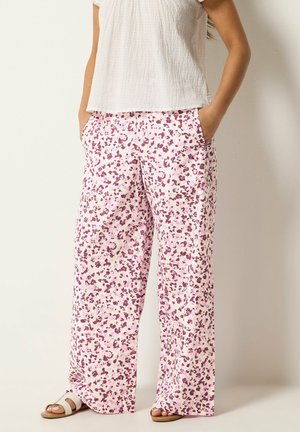 Kobieta nosząca luźne spodnie w różowo-fioletowy wzór z bocznymi kieszeniami, białą teksturowaną bluzkę z krótkim rękawem oraz białe sandały.