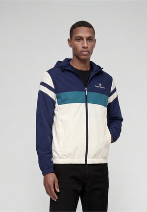 Homme portant une veste zippée bleu marine, sarcelle et blanche avec une capuche et un pantalon noir, debout devant un fond clair uni.