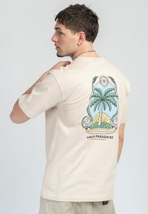 Jeune homme portant un t-shirt beige avec un motif coloré de palmier, serpents et soleil, ainsi que le texte "Virgin Paradise" au dos.