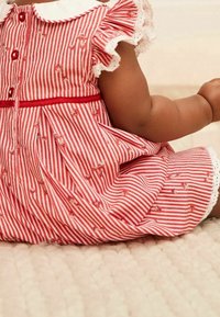 Spædbarn iført en rød og hvid stribet kjole med små blomsterprint og blondekant, der sidder på en blød beige tekstureret overflade.