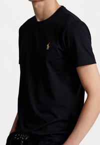 Černé bavlněné tričko s kulatým výstřihem, krátkými rukávy a malým zlatým vyšitým logem na levém prsu. Hladká textura.