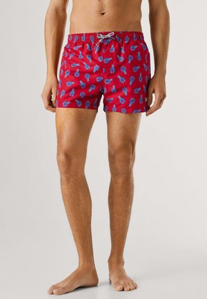 Hombre con pantalones cortos de baño rojos con patrón de langostas azules, de pie descalzo contra un fondo claro y liso.