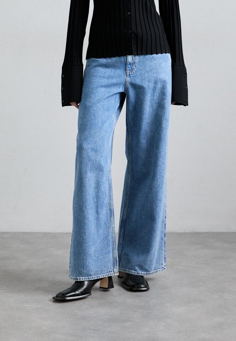 Hellblaue, weit geschnittene Denim-Jeans mit hoher Taille, sichtbaren Nähten und Vordertaschen, kombiniert mit schwarzen, knöchelhohen Stiefeln mit Absatz.
